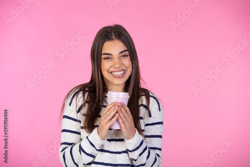 Horizontal shot of pretty brunette woman has long hair, wears warm sweater, holds coffee to go, imagines pleasant moment in life during break, models over pink studio wall