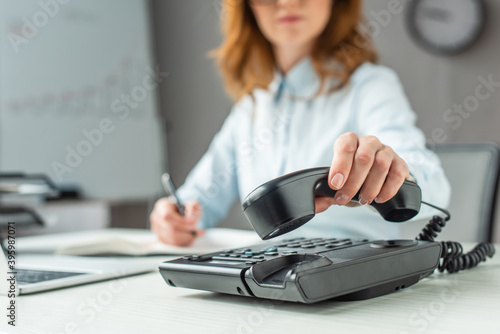 Cropped view of businesswoman putting handset on landline telephone, while sitting at workplace on blurred  photo