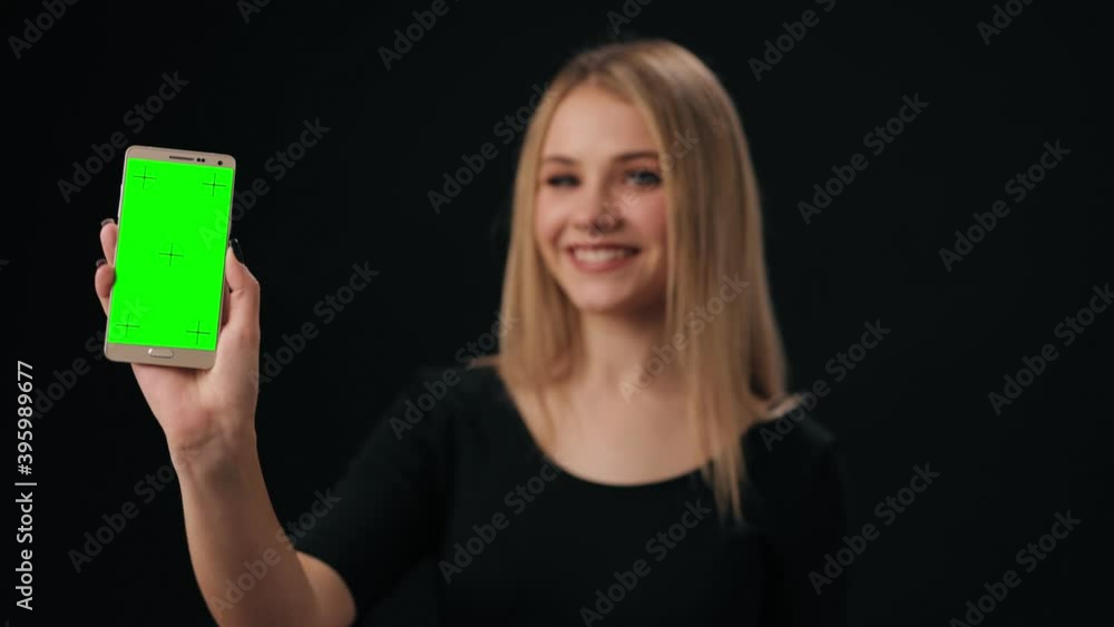 Charming smiling woman in black pullover holding smartphone with green chroma key screen. Isolated over black studio background. Charming smiling woman in black pullover holding smartphone with green chroma key screen. Isolated over black studio background.