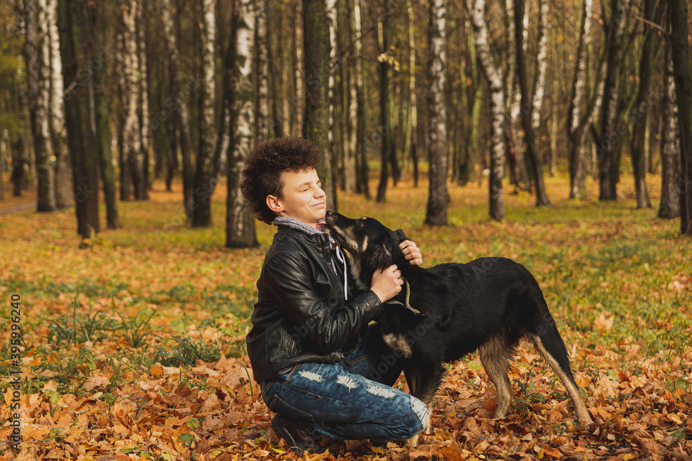 A curly-haired brown-haired boy with a mohawk hairstyle in a leather jacket on the background of an autumn forest plays with a dog.