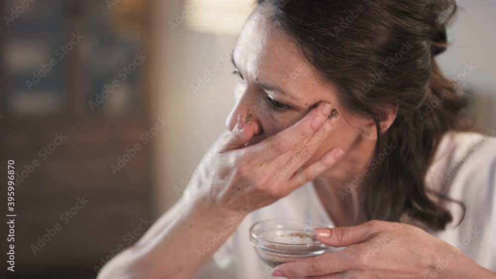 Close-up of middle aged brunette applying scrub made of cream and coffee grounds on face with hands looking at mirror having beauty routine at home Close-up of middle aged brunette applying scrub made of cream and coffee grounds on face with hands looking at mirror having beauty routine at home