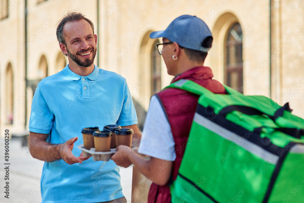 Happy caucasian man customer receiving four cups of coffee from male courier with thermo bag while standing outdoors on the sunny street Happy caucasian man customer receiving four cups of coffee from male courier with thermo bag while standing outdoors on the sunny street