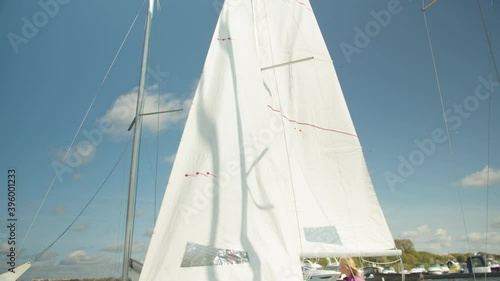 Blonde girl in pink jacket sitting on the stern of a yacht, white sail and yacht blue sky background. Slow motion photo