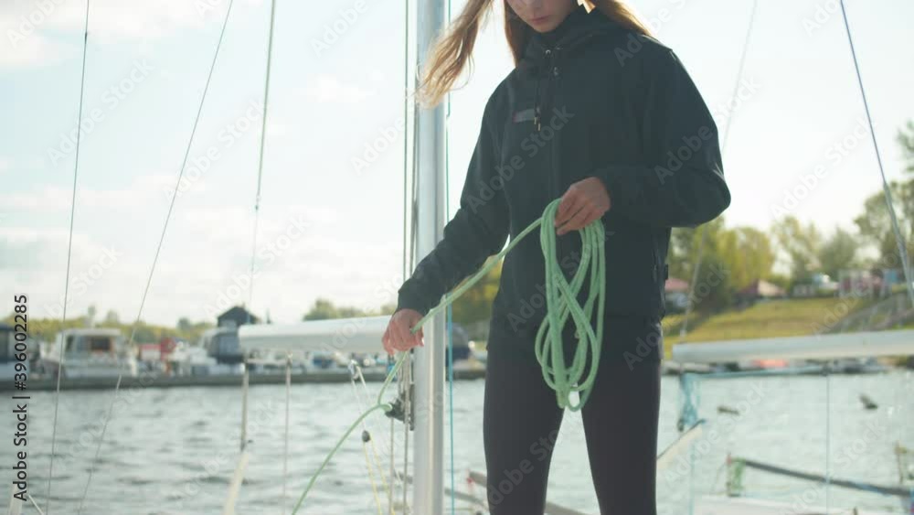 In the dock, a young, pretty sportswoman winds ropes around her arm while pulling a sailing yacht to a regatta on the river In the dock, a young, pretty sportswoman winds ropes around her arm while pulling a sailing yacht to a regatta on the river