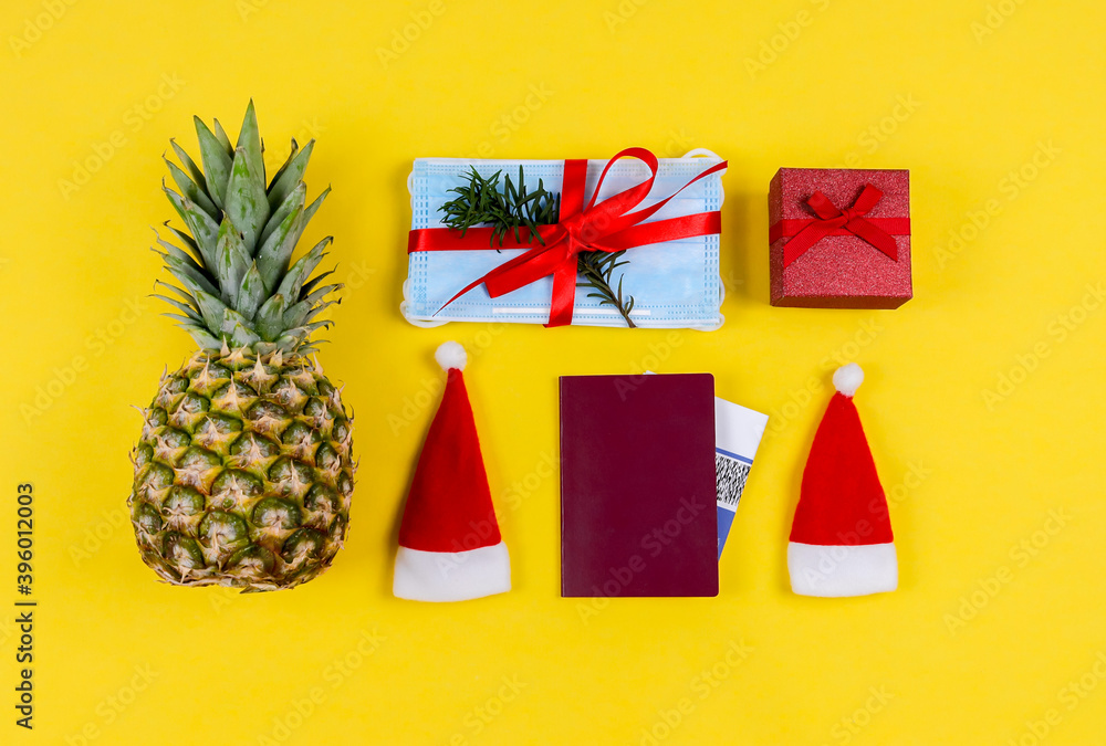 Pineapple, passport and masks.
Pineapple, passport, two hats, santas, gift, ticket and masks with a fir branch are located in the middle on a yellow background, close-up top view.