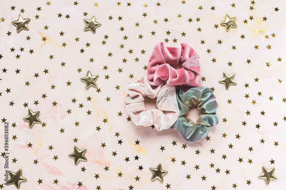 Three trendy velvet pink and holographic shiny metallic scrunchies and golden stars confetti on pink background. Diy accessories and hairstyles for Christmas and New Year party concept, copy space Three trendy velvet pink and holographic shiny metallic scrunchies and golden stars confetti on pink background. Diy accessories and hairstyles for Christmas and New Year party concept, copy space