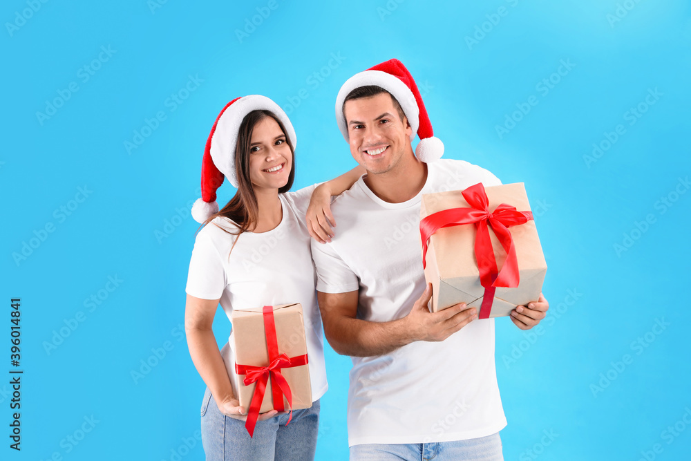 Obraz premium Beautiful happy couple in Santa hats holding Christmas gifts on light blue background