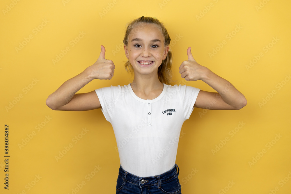 Young beautiful child girl standing over isolated yellow background smiling confident doing the ok signal with her thumbs Young beautiful child girl standing over isolated yellow background smiling confident doing the ok signal with her thumbs