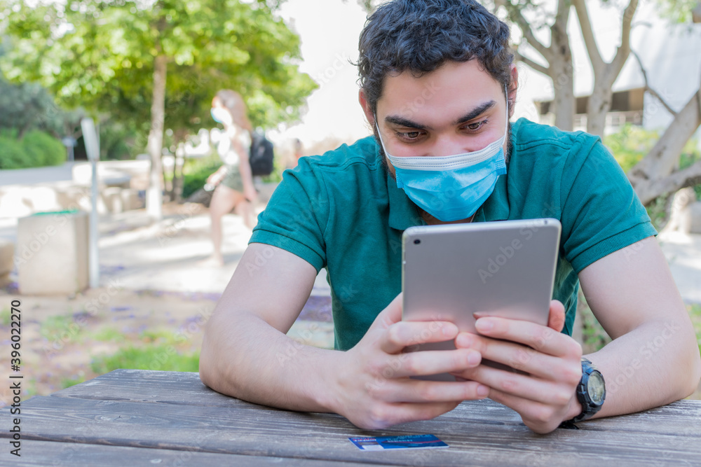 Young man with a protective mask watching multimedia content on a tablet, sitting in a park. Young man with a protective mask watching multimedia content on a tablet, sitting in a park.