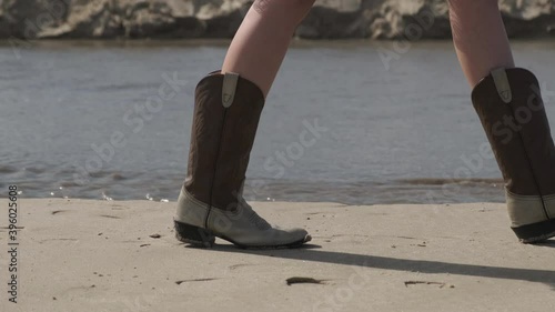 A girl in a white dress and cowboy boots walks on a sandy beach. Focus on your feet.