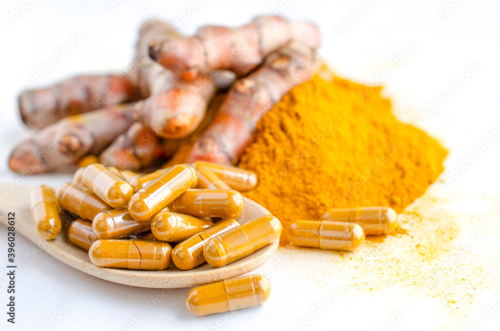 Closed up turmeric or curcumin powder in capsule on wooden spoon, over blur background of fresh turmeric root and powder pile on white background, for health care and natural medical concept