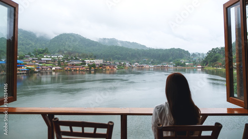 A young woman sitting on balcony and looking at a beautiful lake in mountains village © Farknot Architect