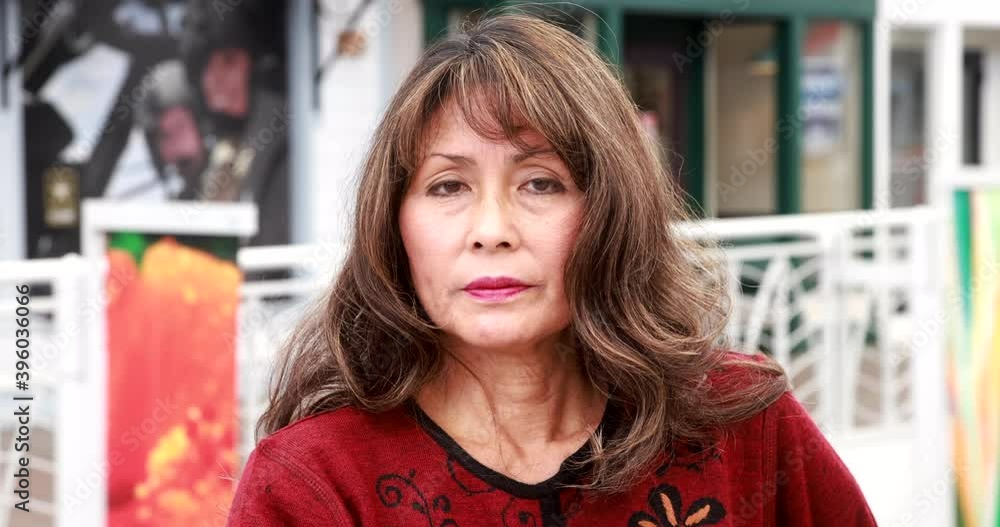 Attractive exotic-looking asian older woman with long curly hair raises her head and looks into camera wearing dark-red coat.  Bright, hand-held medium shot with bokeh background.