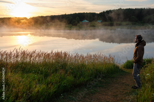 A full-length man stands on the green shore of the lake. Beautiful sunrise, fog over the water.