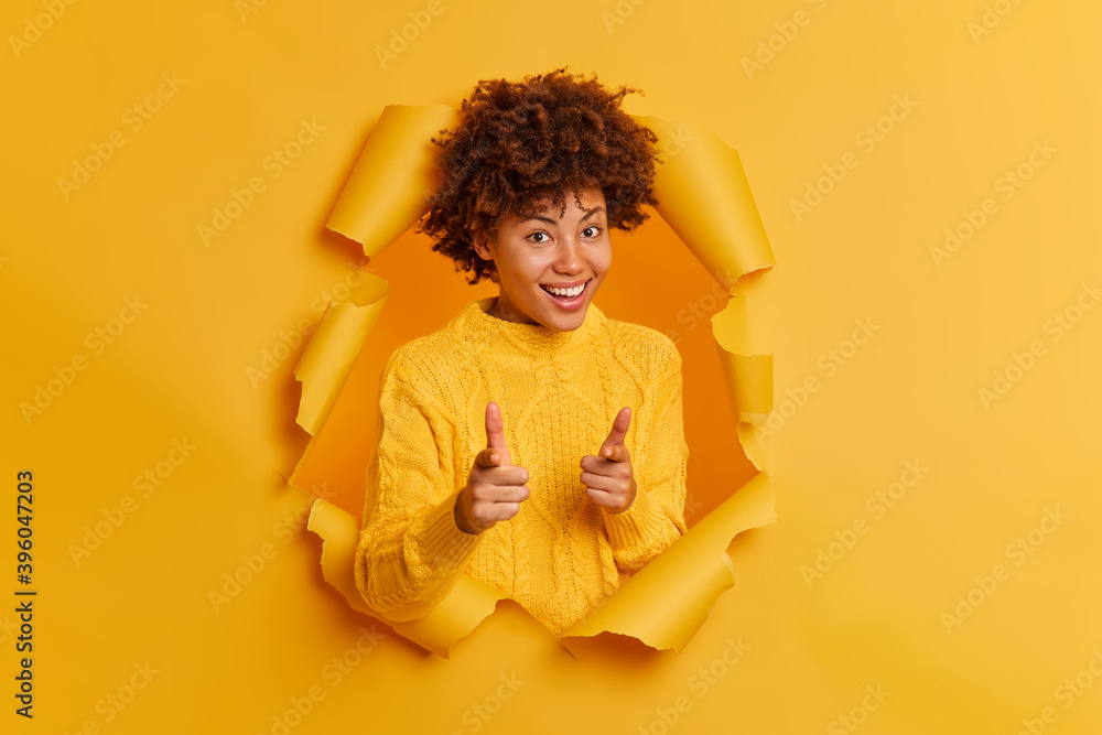 Cheerful dark skinned woman points index fingers at camera picks someone asks to join her giggles positively has curly bushy hair wears casual sweater poses through paper background. Hey you Cheerful dark skinned woman points index fingers at camera picks someone asks to join her giggles positively has curly bushy hair wears casual sweater poses through paper background. Hey you