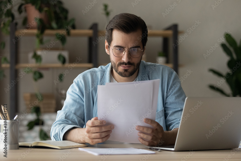 Thoughtful young Caucasian man sit at desk at home office read postal paper letter. Pensive millennial male get message or notice in paperwork or postal correspondence, consider banking document. Thoughtful young Caucasian man sit at desk at home office read postal paper letter. Pensive millennial male get message or notice in paperwork or postal correspondence, consider banking document.