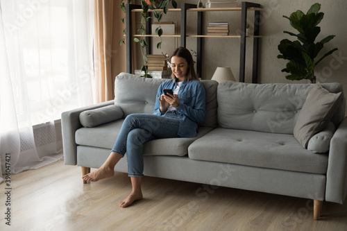 Young Caucasian woman sit relax on cozy couch in living room using modern smartphone gadget. Happy millennial female rest on sofa at home browse surf wireless good internet on cellphone.