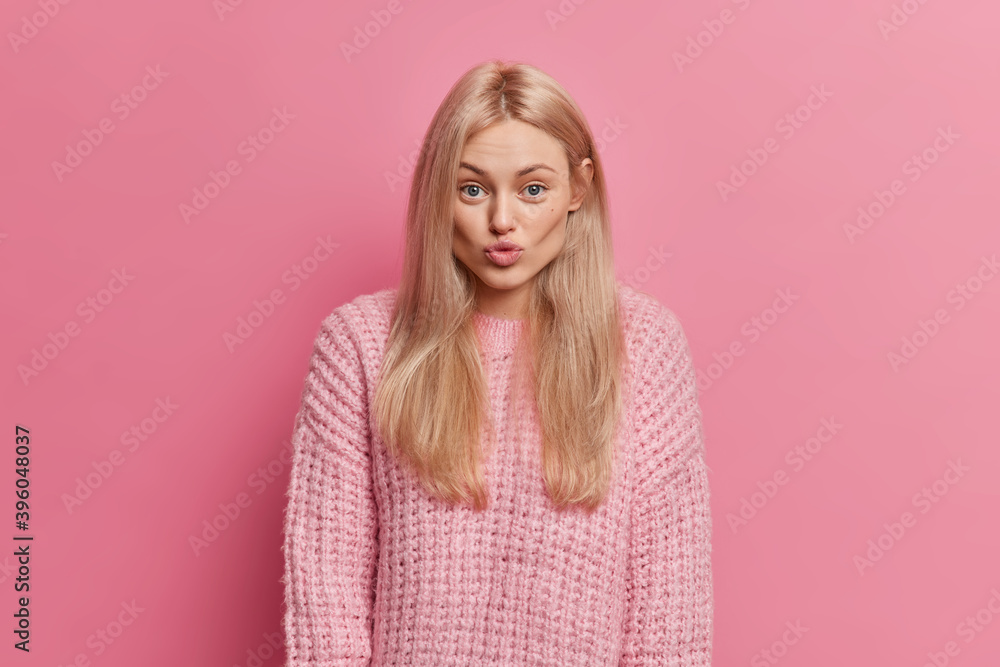 Fototapeta premium Lovely romantic woman with blonde hair keeps lips rounded wants to kiss boyfriend flirts with someone wears warm knitted sweater isolated over pink background. Human face expressions concept