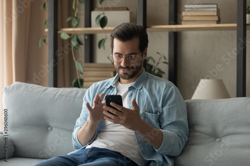 Young Caucasian man in glasses sit rest on sofa at home look at cellphone screen texting or messaging. Millennial male relax on couch in living room browse wireless internet on smartphone gadget.