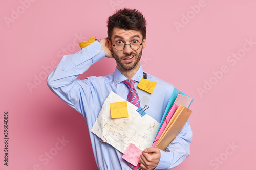 Puzzled office worker does paper work in home office scratches head with embarrassement prepares financial report wears formal clothes stands indoor. Male student hears exam or test results.