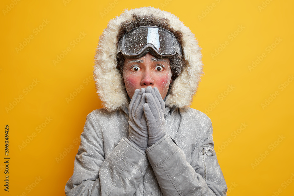Shocked stressed young woman covers mouth with hands wears hooded jacket and gloves covered with hoarfrost feels cold during seasonal outdoor activities in Scandinavia isolated over yellow background Shocked stressed young woman covers mouth with hands wears hooded jacket and gloves covered with hoarfrost feels cold during seasonal outdoor activities in Scandinavia isolated over yellow background