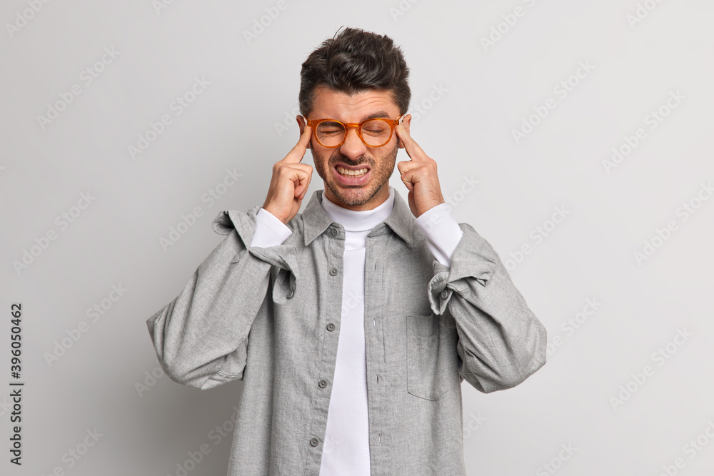 Stressed European unshaven man suffers headache keeps hands on temples clenches teeth and tries to relieve pain wears grey shirt and spectacles poses indoor. People and health problems concept Stressed European unshaven man suffers headache keeps hands on temples clenches teeth and tries to relieve pain wears grey shirt and spectacles poses indoor. People and health problems concept