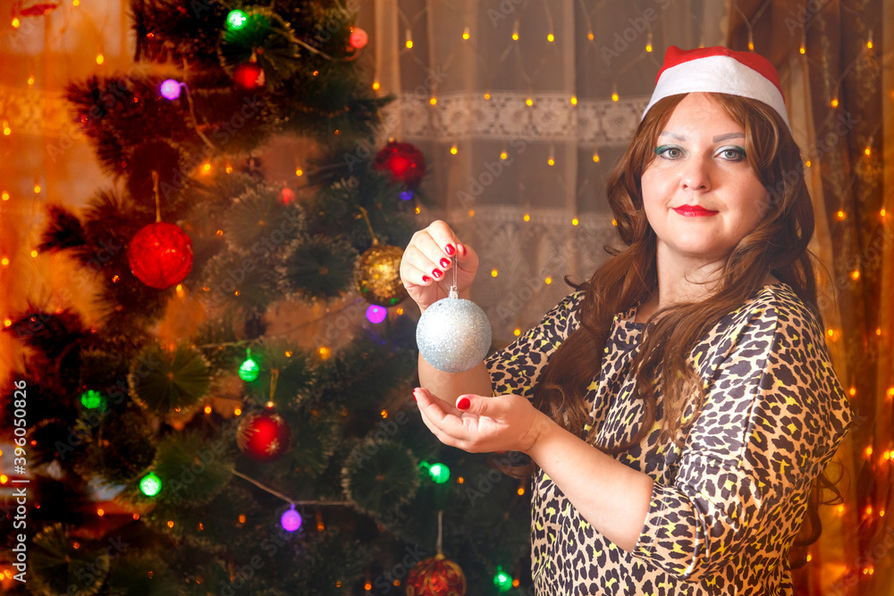 Naklejka premium A brown-haired woman in a Santa hat by a Christmas tree with a decoration ball in her hands.