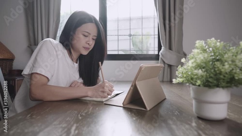 Young asian girl working on final exam revision, using tablet for online class lesson, at home schooling, use pencil write down on small notebook, job searching opportunity, sitdown alone at home desk photo