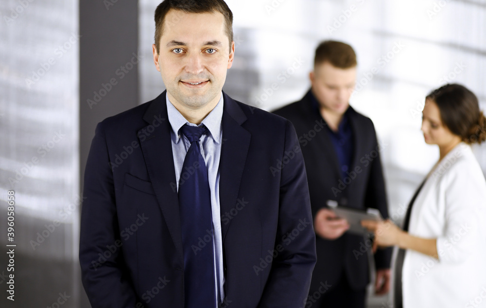 Portrait of a self-confident middle aged businessman in a blue suit, standing in a modern office with his colleagues at the background. Concept of business success Portrait of a self-confident middle aged businessman in a blue suit, standing in a modern office with his colleagues at the background. Concept of business success