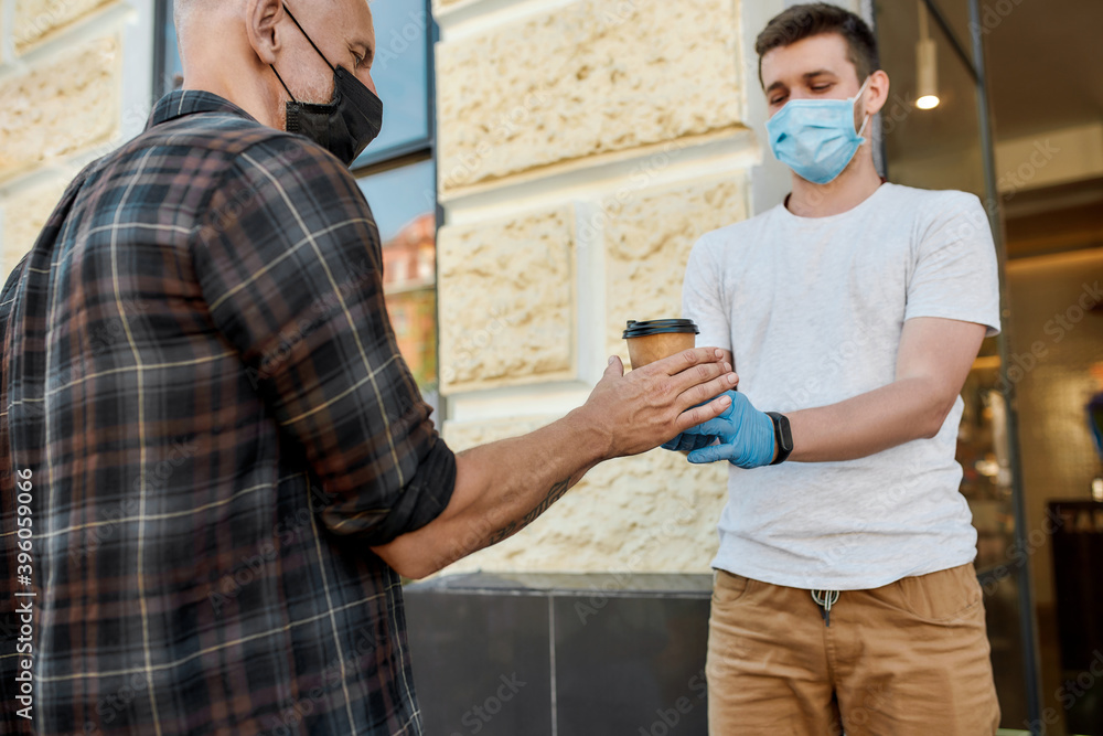Middle aged man wearing mask receiving hot coffee drink from hands of cafe assistant in protective gloves while collecting his takeaway order during coronavirus lockdown Middle aged man wearing mask receiving hot coffee drink from hands of cafe assistant in protective gloves while collecting his takeaway order during coronavirus lockdown