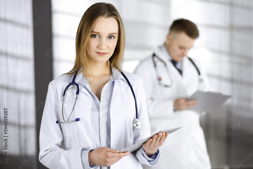 Professional woman doctor with a stethoscope is using a computer tablet, while she is standing together with her colleague in a clinic. Young doctors at work in a hospital. Medicine and healthcare Professional woman doctor with a stethoscope is using a computer tablet, while she is standing together with her colleague in a clinic. Young doctors at work in a hospital. Medicine and healthcare