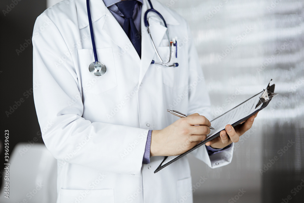 Unknown male doctor standing and working with clipboard of medication history records in clinic at his working place, closeup. Perfect medical service in hospital. Medicine and healthcare concept Unknown male doctor standing and working with clipboard of medication history records in clinic at his working place, closeup. Perfect medical service in hospital. Medicine and healthcare concept