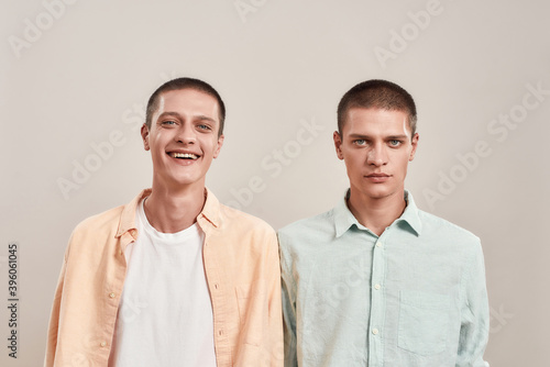 Portrait of two young caucasian men, twin brothers looking at camera with different face expressions while posing together isolated over beige background