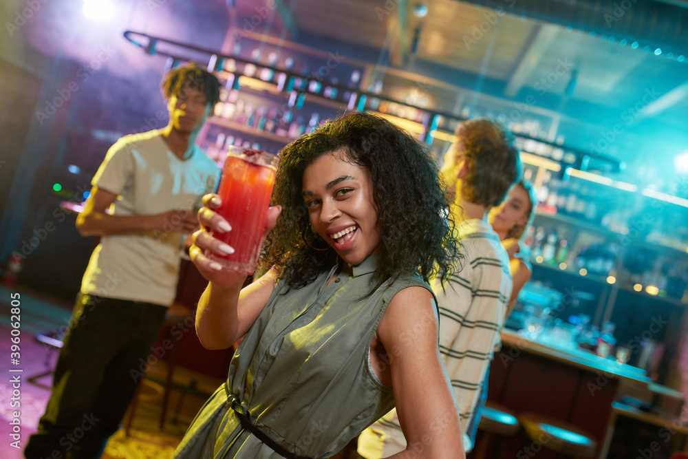 Attractive mixed race young woman looking at camera while posing with a cocktail in her hand and friends chatting, having drinks at the bar counter in the background Attractive mixed race young woman looking at camera while posing with a cocktail in her hand and friends chatting, having drinks at the bar counter in the background
