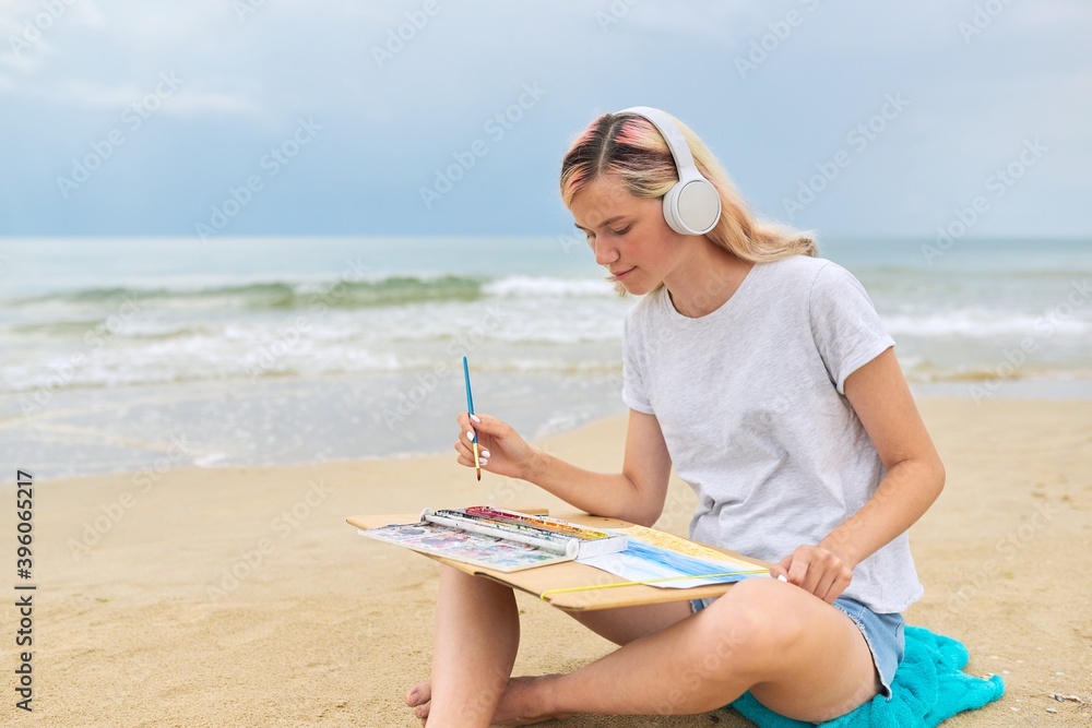 Young woman teenager in headphones drawing sketch with watercolors sitting on sea beach