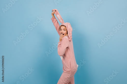 Surprised girl in rabbit costume holding ears on blue background. Studio shot of pretty young woman wears bunny kigurumi.