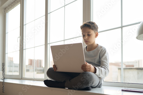 10 years old boy sitting on sill of big white window and pushing button on laptop, sunlight. High quality photo