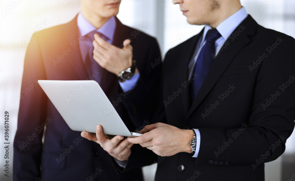 Business people discussing a presentation at meeting, standing in a sunny modern office. Unknown businessman with a colleague search for some information at the laptop, lawyers at negotiation