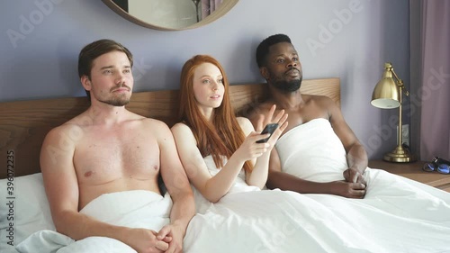Diverse trio watches TV in the bedroom. Concept of free relationships. Red-haired woman sitting with two men in bed photo