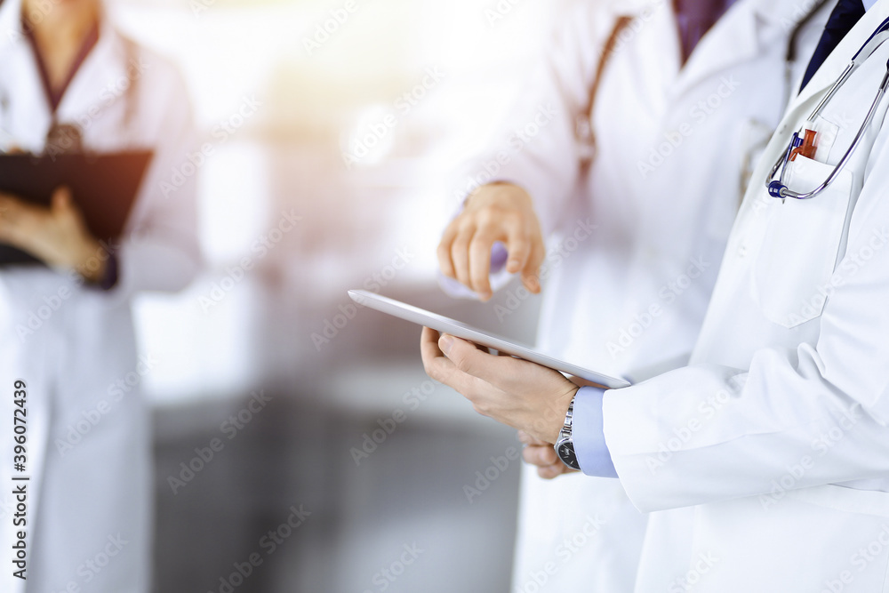 Group of unknown doctors use a computer tablet to check up some medical names records, while standing in a sunny hospital office. Physicians ready to examine and help patients. Medical help, insurance Group of unknown doctors use a computer tablet to check up some medical names records, while standing in a sunny hospital office. Physicians ready to examine and help patients. Medical help, insurance