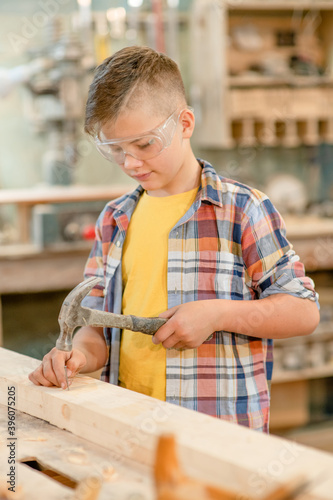 Young boy hammering a nail in wooden plank in carpentry workshop