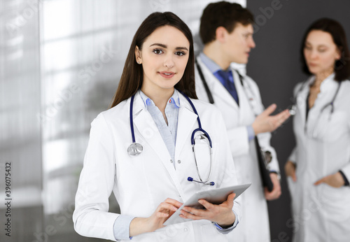 Smiling intelligent woman-doctor is holding a tablet computer in her hands, while she is standing together with her colleagues in a clinic. Physicians at work. Perfect medical service in a hospital