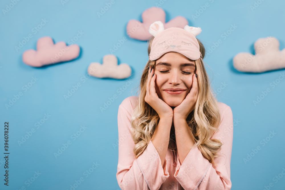 Excited girl in sleep mask smiling with eyes closed. Studio shot of blithesome woman in pajama posing on blue background with clouds. Excited girl in sleep mask smiling with eyes closed. Studio shot of blithesome woman in pajama posing on blue background with clouds.