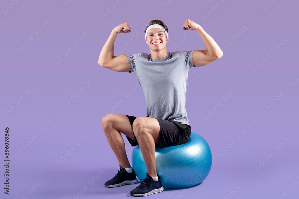 Positive young sportsman sitting on fitness ball and showing his strength on lilac studio background Positive young sportsman sitting on fitness ball and showing his strength on lilac studio background