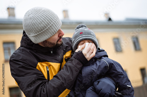 a man helping to blow nose of his little son wearing winter jacket and knit hat