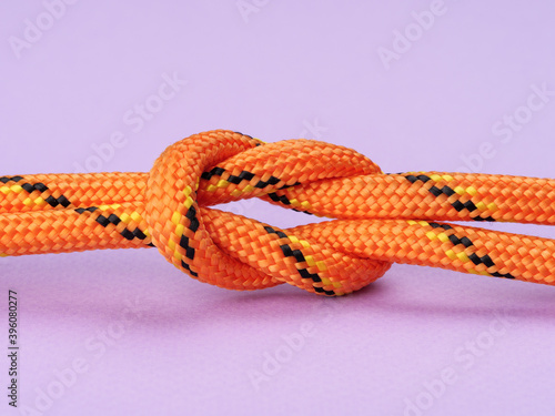 A square knot made of nylon orange rope on a pastel purple background.