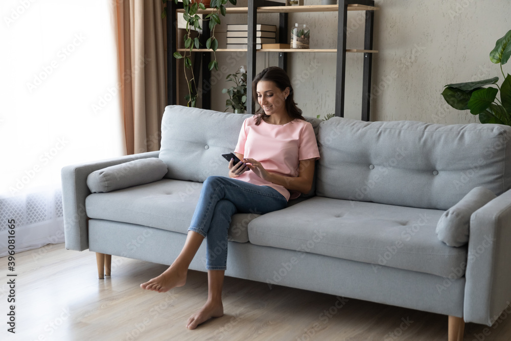 Full length happy relaxed barefoot woman sitting on cozy sofa, involved in distant mobile communication, web surfing information, shopping in online store, using telephone applications at home.