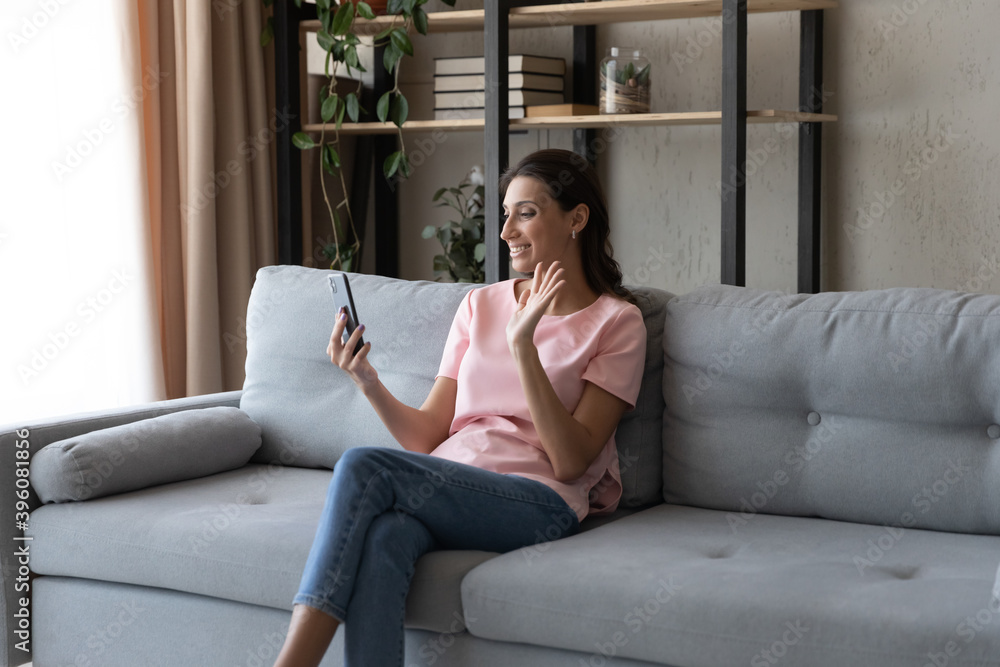 Fototapeta premium Relaxed young happy arabic indian mixed race woman sitting on sofa, involved in video call pleasant conversation using modern technology cellphone application, looking at web camera at home.