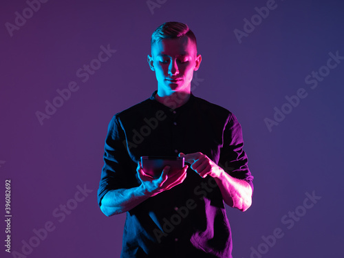 Man with a tablet on a purple background. It is illuminated by neon light. Concept - he uses a tablet application. Guy is using the app in the gadget. Student is using some kind of application