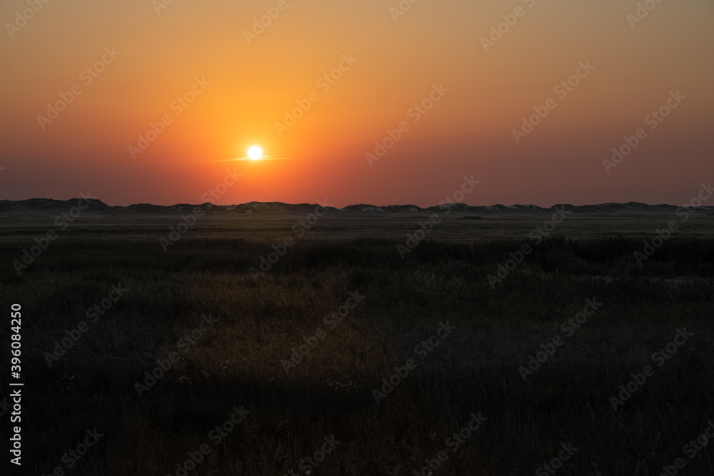 Wide panoramic view over the grassland between the dike and the beach against the deep setting sun.